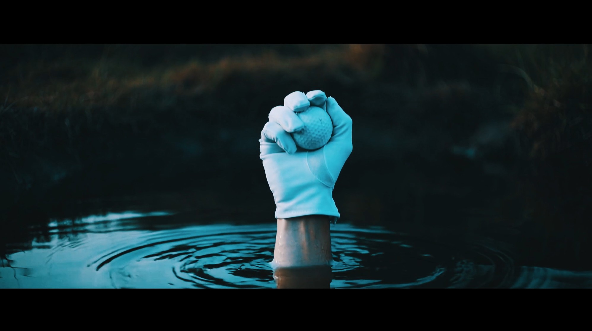 A white-gloved hand grips a golf ball while rising from dark water